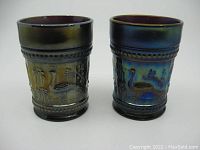 Two amethyst purple carnival glass tumblers with embossed peacock and fountain pattern, front view showing detailed design.