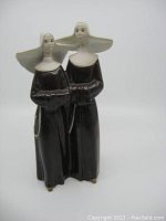 Front view of the porcelain figurine showing two standing nuns dressed in black habits and white wide head coverings, one holding rosary with cross.