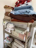 Full view of plastic shelves loaded with assorted bedding, blankets and sheets including new packaged sheets at bottom shelf and folded blankets on upper shelves.