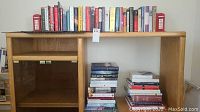 Wide shot of a wooden shelf with a row of books on the top shelf and multiple stacked books on the floor beneath
