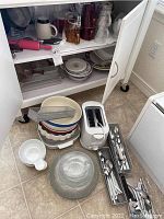 Various ceramic plates, bowls, clear glass dish, white toaster, metal utensil trays with white-handled cutlery and spoons on floor