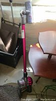 Dyson V6 cordless vacuum cleaner standing upright on a floor next to furniture with visible dustbin and pink wand.
