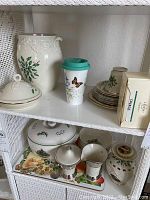 Two shelves showing a variety of Lenox holiday dishware, including a large pitcher, covered sugar bowl, plates and cups with holly decoration. Also includes a travel tumbler with butterfly motif.