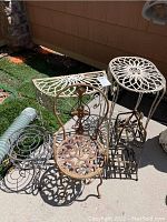 Five metal plant stands grouped together on a patio, showing variety of styles and finishes including white and rustic bronze metals.