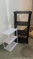 Photo showing black four-tier plastic shelving unit next to white three-tier plastic shelving unit in basement setting.