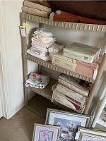 Shelf unit holding folded white towels, packaged bedding, and blankets stacked on three levels with some artwork below on the floor.