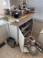 Front angle view showing white wooden cart with butcher block top, drawer, double door cabinet open to reveal storage with pots and pans, open lower shelf with mixing bowls, items on top not included.