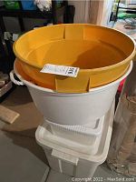 White rectangular plastic storage tub with lid, large white round bucket with rope handles, and large yellow plastic basket stacked inside one another