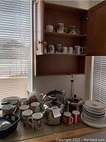 Image showing various ceramic mugs, teacups on shelves and countertop, Corelle dishes stacked on side, stainless steel kettles and pots, plus small pans and utensils.