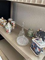 Photo showing a variety of decorative ceramic teapots and clear glass/crystal pieces arranged on a shelf.