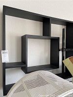 Photo showing front and partial side view of the black geometric wall shelf mounted on a white wall with interconnected rectangular compartments.