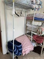 Full side view of the white metal clothing rack with several folded blankets on shelves and hangers on the rod.
