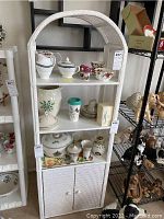 Front view of the white wicker storage cabinet showing three shelves and bottom doors, with various items on shelves not included in sale.
