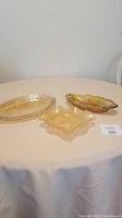 Photo showing three amber carnival glass trays and dishes on table with cloth, including oval, square, and boat-shaped dishes