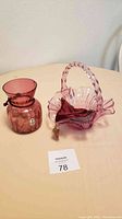 Photo showing the cranberry glass vase and cranberry glass basket side by side on a table with lot number card.