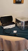 Hamilton Beach electric grill and large roasting pan placed on a table, grill lid open, roasting pan stacked one over the other beside grill.