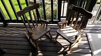 Two small wooden children's rocking chairs positioned side by side on a wooden floor, showing overall weathered condition and design.