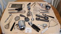 Photo showing various metal and plastic kitchen utensils laid out on table including hand mixer, cheese grater, sifter, measuring spoons, ice cream scoopers, wire whisk, potato masher, and other scoops and cutters.