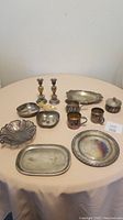 Full view of the 9 silver items on round table showing plates, trays, candleholders, cups, and small pots