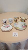 Five fine china items on a beige tablecloth include two creamers, a sugar bowl, a candy dish, and a small cup with pink rose floral designs and gold rims.