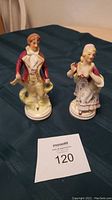 Photo showing both porcelain figurines of a man and woman, placed on a dark cloth background with a Maxsold lot tag labeled 120.