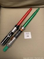 Two Star Wars lightsabers, one with a red blade and one with a green blade, shown side by side on a beige surface.