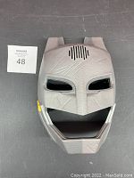 Front angle view of gray Batman mask showing textured detail, eye openings, speaker grill, and yellow side button.
