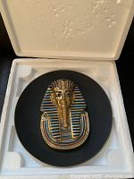 Collector plate with detailed 3D golden mask design inside white foam protective case, showing top view of the plate and mask.