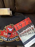 Photo showing the black sweatshirt with Team Canada 1972 embroidered logo and the commemorative book cover.