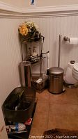 Photo showing metal stand with soap dispenser, artificial flowers, trash can, wall-mounted toilet paper holder, and grey organizer basket on floor corner of bathroom