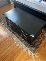 Image of the entire black wooden chest on an oriental rug, showing the smooth top and front drawers with handles.