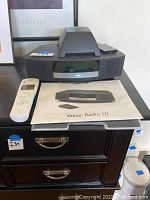Bose Wave Radio III main unit on drawer with user manual and black remote on top, white remote in front