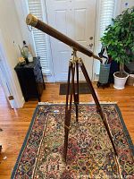 Full view of the brown telescope mounted on a wooden tripod in an indoor room with hardwood floors and oriental rug.