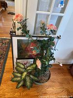 Three artificial flower decorations including pink roses and large green leaves arranged beside two framed floral pictures leaning against a window and a small flashlight.