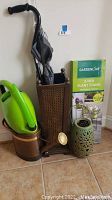 Overall view of the lot showing two watering cans (green plastic and copper metal), the tall brown umbrella holder, boxed Gardenline 3-tier plant stand, and green ceramic lantern.