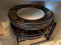 Stack of large black china plates with gold Forbidden City designs stacked on a black metal rack.