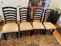 Four matching dark wood dining chairs with ladder backs and beige upholstered seats placed in a row on a patterned rug in a living room.