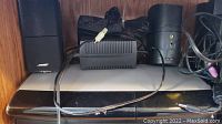 Close-up of two Bose satellite speakers, power adapter, and cables on a wooden shelf.
