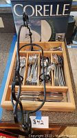Drawer with neatly arranged stainless steel flatware forks, knives, spoons, and teaspoons in wooden tray compartments, Corelle dish box visible behind