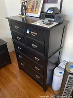Front angled view of black chest of drawers showing 7 drawers with silver metal handles and some items on top.