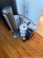 Overview of the lot showing the two Lasko tower fans (one out of box, one boxed), two white decorative containers with lids, small trash can with trash bags, and a slow close toilet seat.