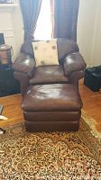 Brown leather Lazyboy recliner chair and matching ottoman with a light beige decorative pillow placed on the chair. Chair and ottoman are on a patterned rug with wooden flooring.