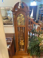 Full view of tall wooden grandfather clock standing next to stairs and piano.