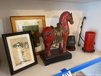 Wide view photo showing two framed Asian style paintings, red stallion horse statue, owl figurine, red candle, and candle holder on a shelf