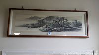 Framed Asian sea to mountain landscape artwork hung on a wall, showing the full piece in a wooden frame with glass.