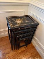 Stacked set of four black nesting tables with floral and bird designs on tabletops against a white wall on wooden floor.