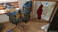 Overview photo showing both stained glass pieces on floor near window light, butterfly with wire embellishments on left, cardinal framed panel on right.