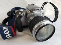 Front angled view of the silver Canon EOS Rebel T2 film camera with large zoom lens and Canon branded strap
