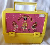 Front view of yellow plastic lunchbox with Cherry Merry Muffin design panel showing three girls and polka dot background, handle up, two latch closures on bottom front