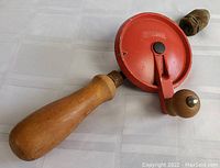 Side view of the hand drill showing the wooden handle and red metal crank wheel with wooden knob.